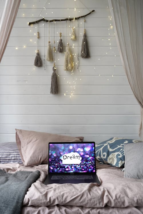 Free Serene bedroom interior with a laptop displaying 'Dream', decorated with soft pillows and warm lights. Stock Photo