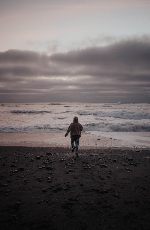 Free Back view of anonymous person in warm clothes running towards waving sea against cloudy sky in evening Stock Photo